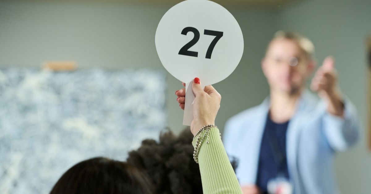 Close-up of a hand holding a paddle with number 27 as the auctioneer gestures in the background